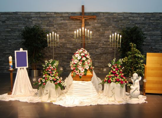 Trauerhalle mit Sarg, Blumenschmuck und Kerzenleuchtern vor einem Holzkreuz