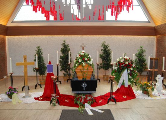 Trauerhalle mit Sarg auf rotem Tuch, Kerzen und weißen Schleifen