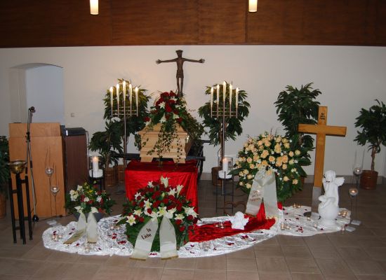 Trauerhalle mit Sarg, weißen Rosen und rotem Tuch vor einem Kreuz