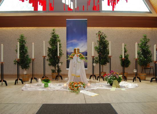 Urne auf hellem Tuch vor einem Bild mit Himmelmotiv, daneben Kerzen und Blumen